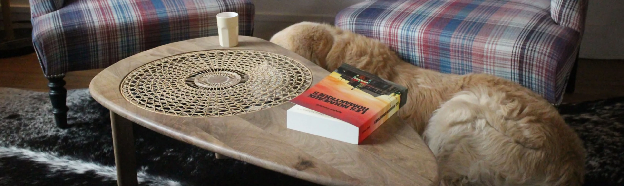 Photo d'ambiance d'une table basse réaliser par Inès Hayaud en noyer avec un cannage soleil. on voit la table dans un salon avec des fauteuils, un livre, un café, un chien.